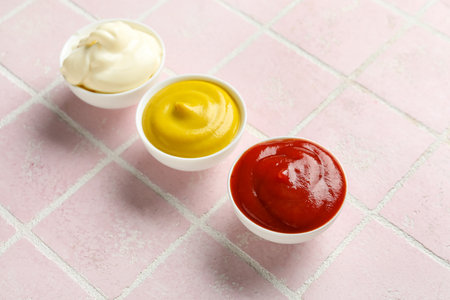 Bowls of ketchup, mayonnaise and mustard on pink tiled table, closeupの写真素材