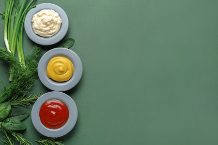 Plates of ketchup, mayonnaise and mustard with herbs on green backgroundの写真素材