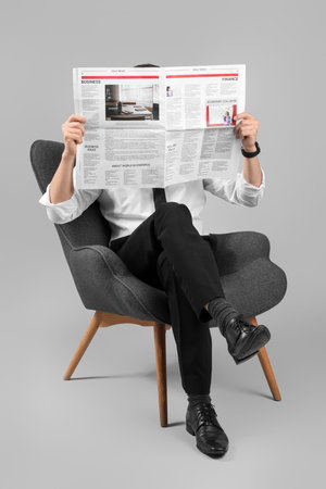 Handsome businessman reading newspaper in armchair on gray backgroundの写真素材