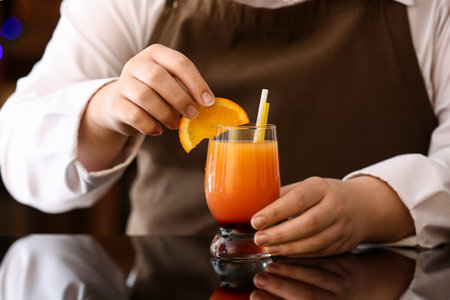 Female bartender placing orange slice in glass of tasty Tequila Sunrise on dark table in barの写真素材