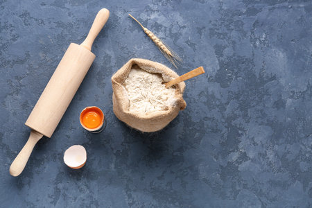 Sack bag of wheat flour, rolling pin and cracked egg on blue grunge background