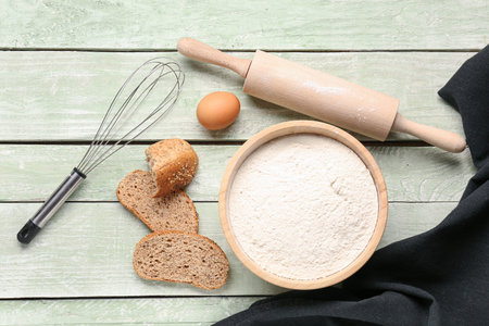 Bowl with wheat flour, bread, egg and utensils on green wooden table