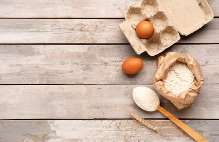 Composition with paper bag of wheat flour, spoon and eggs on gray wooden table
