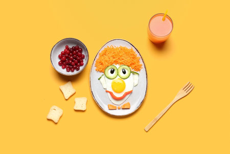 Plate with funny breakfast in shape of clown, berries and glass of juice on orange background