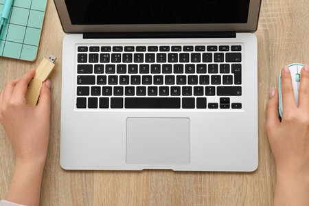Female hands with modern laptop, USB flash drive and computer mouse on wooden background, closeup
