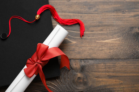 Diploma with red ribbon and graduation hat on wooden tableの写真素材