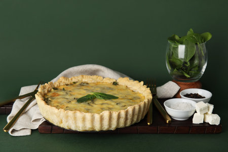Board with delicious quiche, spinach leaves, feta cheese and seasoning on green background
