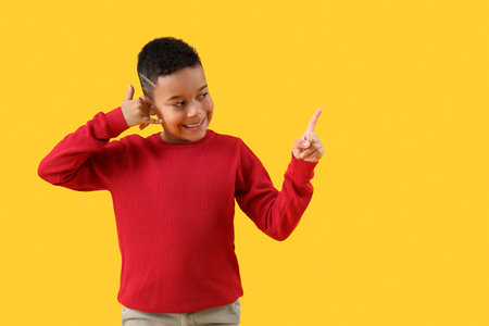 Little African-American boy showing "call me" gesture and pointing at something on yellow backgroundの写真素材