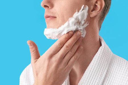 Young man applying shaving foam onto face against light blue background, closeupの写真素材