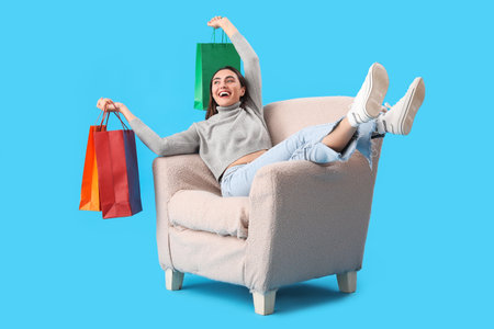 Young woman with shopping bags sitting in soft armchair on blue backgroundの写真素材