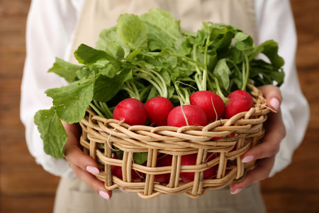 Woman holding wicker basket of ripe radish with green leaves on wooden background, closeupの写真素材