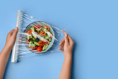 Female hands wrapping bowl with salad by stretch wrap on blue backgroundの写真素材