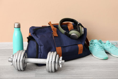 Sports bag with headphones, sneakers, dumbbell and bottle near beige wall in gymの写真素材