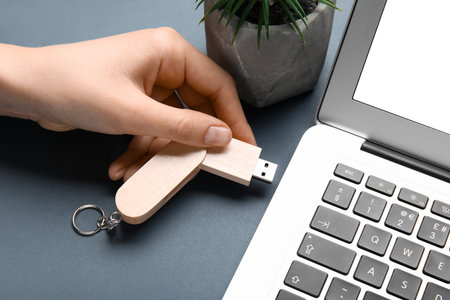 Woman plugging wooden USB flash drive into modern laptop on gray table