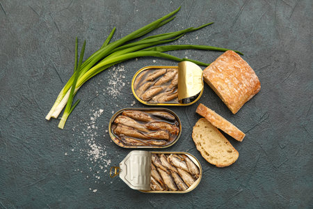 Opened tin cans with sprats in oil, green onion and bread on black tableの写真素材