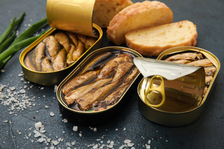 Opened tin cans with sprats in oil and bread on black tableの写真素材