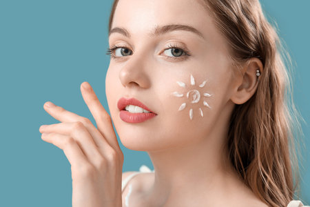 Beautiful young woman with sunscreen cream on her face against blue background