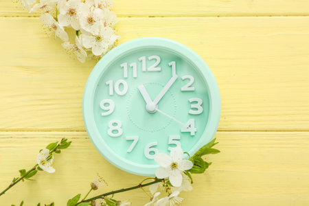 Blooming spring branches and blue clock on yellow wooden background