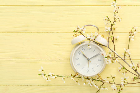 Blooming spring branches and white alarm clock on yellow wooden background