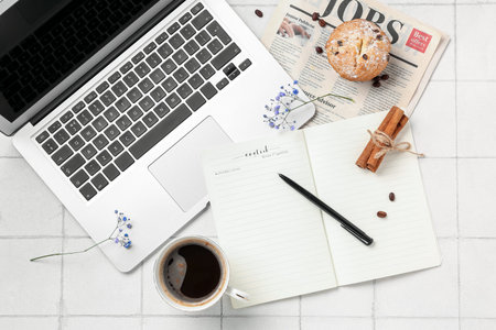 Cup of coffee with notebook, flowers, laptop, muffin and newspaper on white tile background