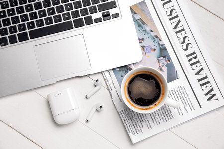 Cup of coffee with earphones, newspaper and laptop on white wooden background