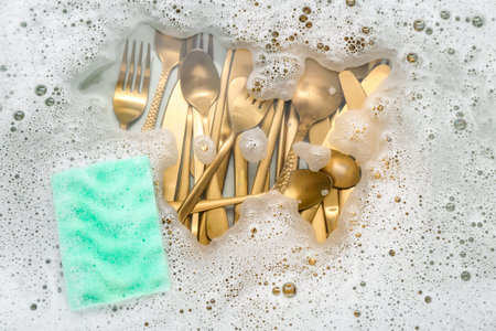 Cleaning sponge with golden cutlery and foam in water, closeup