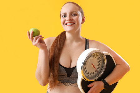 Young overweight woman with scales and apple on yellow background