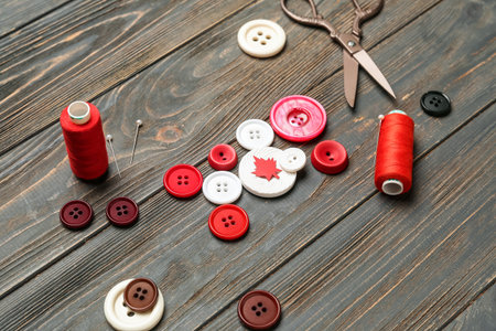 Buttons with maple leaf, thread spools and scissors on dark wooden background, closeup. Canadian...