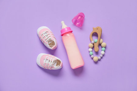 Bottle of milk for baby with rattle and booties on lilac backgroundの写真素材