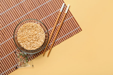 Raw instant noodles in bowl with chopsticks, seasoning and bamboo mat on yellow backgroundの写真素材