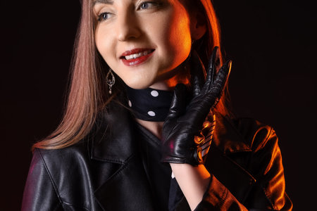 Beautiful young woman in leather gloves and jacket on dark background
