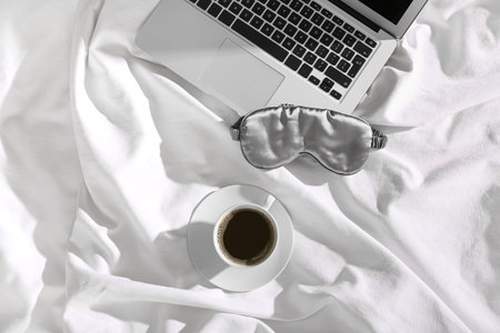 Sleeping mask, laptop and cup of coffee on bedの写真素材