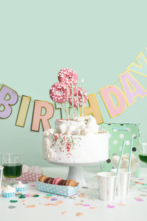 Birthday cake with different sweets on table near green wall