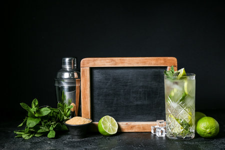 Chalkboard, shaker, glass of cold mojito and ingredients on dark backgroundの写真素材