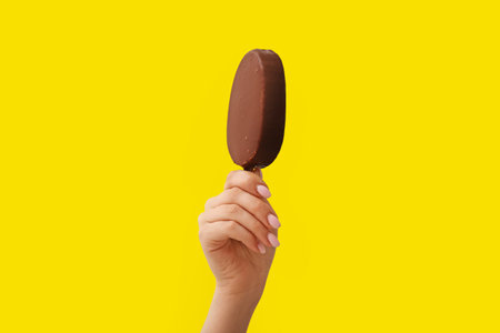 Woman holding delicious chocolate covered ice cream on stick against yellow backgroundの写真素材