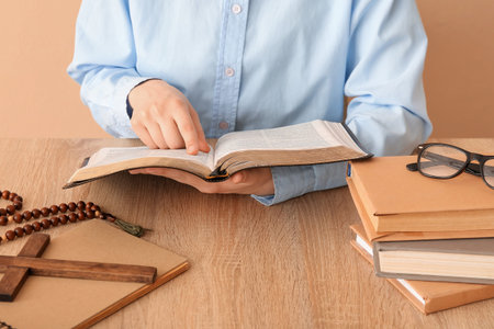 Woman reading Holy Bible at table on beige background, closeup