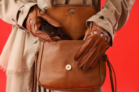 Beautiful young woman in leather gloves and coat putting sunglasses in bag on red background,...
