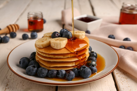 Pouring honey onto tasty pancakes with banana and blueberry on light wooden background, closeupの写真素材