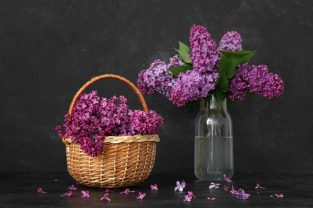 Wicker basket and glass vase of beautiful lilac flowers on table near grunge black wallの写真素材