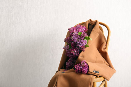 Stylish female coat with beautiful lilac flowers on chair near light wallの写真素材