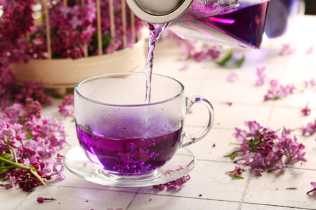Pouring of hot lilac tea into glass cup and flowers on light tile table, closeupの写真素材