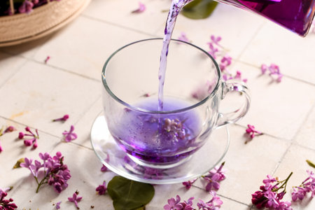 Pouring of hot lilac tea into glass cup and flowers on light tile table, closeupの写真素材