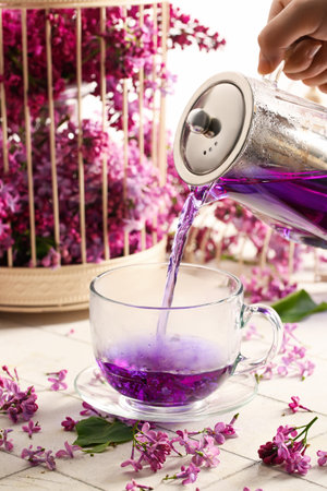 Woman pouring of hot lilac tea into glass cup and flowers on light tile table, closeupの写真素材