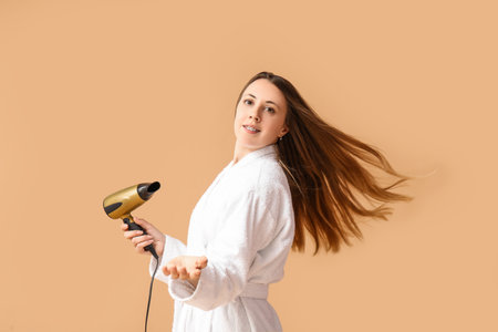 Beautiful young woman in bathrobe and with hair dryer on beige backgroundの写真素材