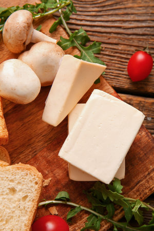 Board with processed cheese, bread and vegetables on brown wooden background