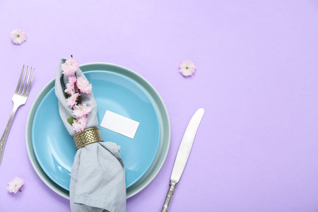 Beautiful table serving with cutlery, blooming tree branch, flowers and folded napkin on lilac backgroundの写真素材
