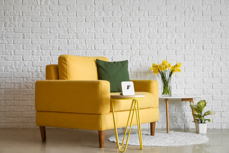 Cozy yellow armchair and vase with blooming narcissus flowers on coffee table near white brick wallの写真素材