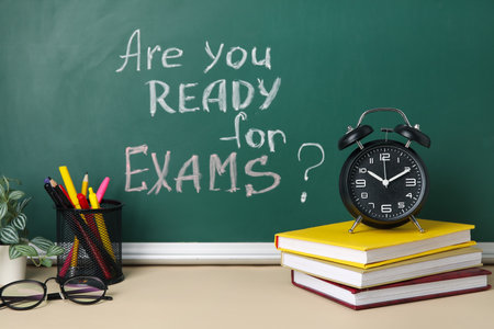 Alarm clock, books and pen holder on table against blackboard with question ARE YOU READY FOR EXAMS?の写真素材