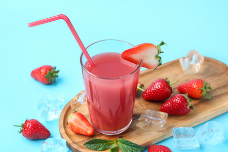 Board with glass of tasty strawberry juice and ice cubes on blue backgroundの写真素材