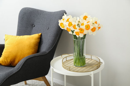 Cozy gray armchair and vase with blooming narcissus flowers on coffee table near white wallの写真素材